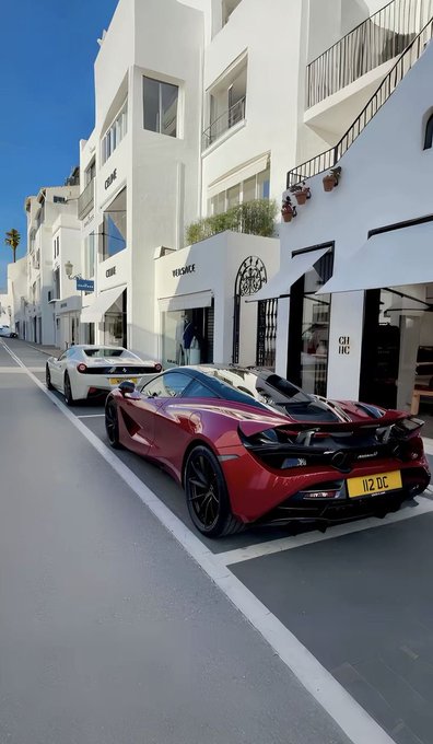 A red McLaren supercar with the license plate '112 DC' is parked on a city street in front of white buildings, including a storefront labeled 'CHIC'.