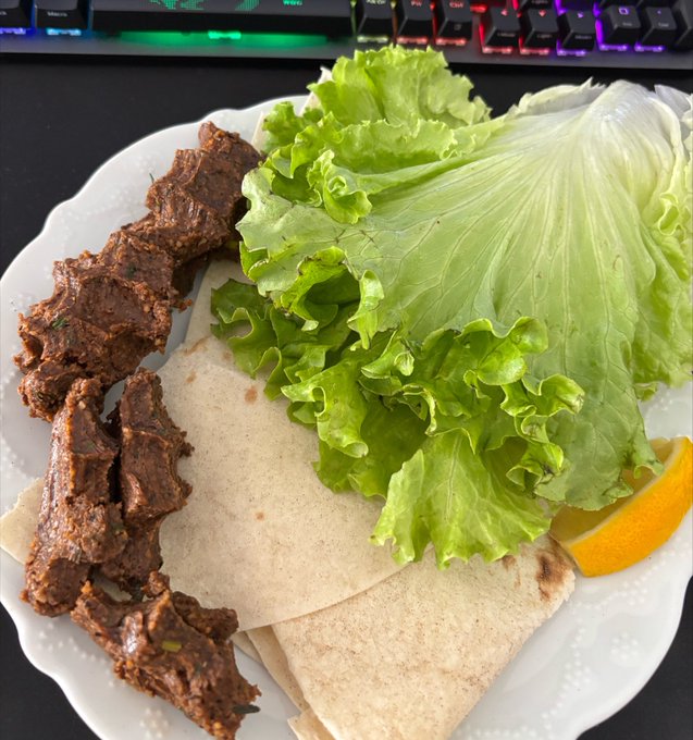 A plate of food containing grilled meat, flatbread, lettuce, and a lemon wedge, with a backlit RGB keyboard visible in the background.