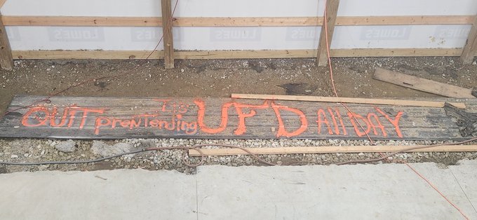 A wooden plank on a construction site with orange spray-painted text reading 'QUIT pretending' and 'UP.D ADAY' lying on dirt near a concrete foundation.