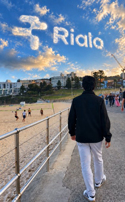 A person with long dark hair wearing a black jacket and white pants walks along a beachside path, with the text 'Ridia' displayed in cloud-writing in the sky above the ocean.