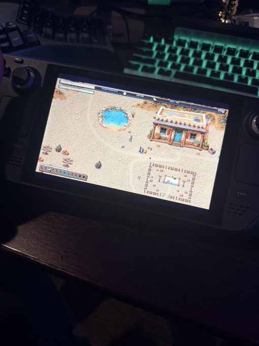 A Steam Deck displays a top-down pixel art game featuring a desert town with a saloon, pond, and characters, set on a dark desk beside an RGB mechanical keyboard. The image visually supports the post's humorous claim of having developed a "Stardew Valley clone" called "Mountaindew Canyon."