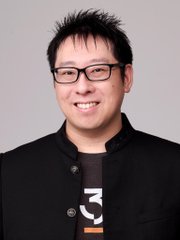 A headshot of an Asian man with glasses and spiky hair smiling, wearing a black jacket with a white graphic design on the shirt underneath.