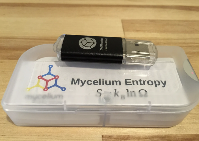 This image shows a Mycelium Entropy device, a USB drive designed for generating truly random numbers, alongside its packaging which bears the name and logo. The device is a black USB stick with "Get Mycelium Bitcoin Wallet" printed on it.