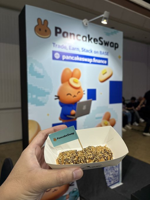 A hand holding a paper food container filled with a pastry topped with seeds, with a small 'PancakeSwap' flag in it, taken in front of a PancakeSwap promotional banner at a trade event.