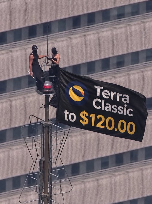 Two masked activists unfurl a "Terra Classic to $120.00" banner from atop a streetlight, rallying behind the $LUNC meme campaign pushing for a dramatic price surge.