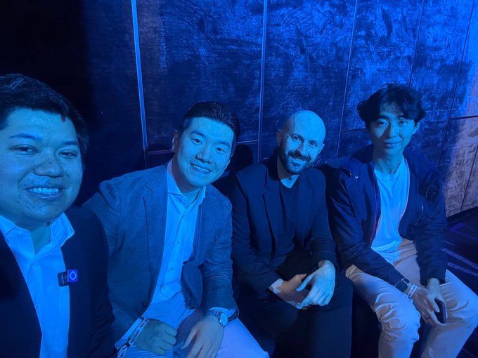 Four men sit closely together in a dimly lit venue bathed in blue light, posing for a selfie-style photo.