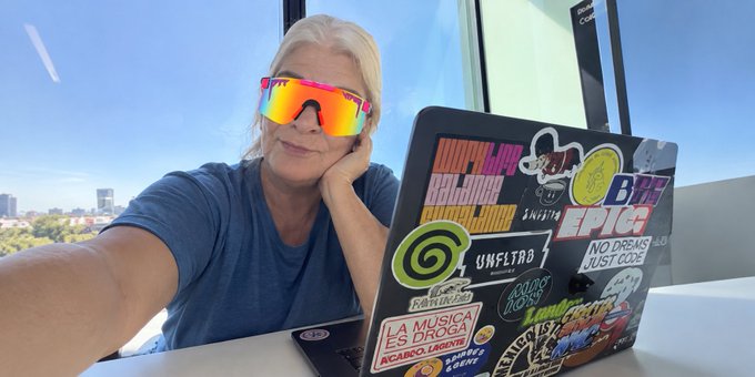 A blonde woman wearing reflective orange-pink sunglasses rests her chin on her hand while posing with a laptop covered in colorful stickers, including 'La Música Es Droga,' 'Unfiltered,' and 'Epic,' against a window with a city view.