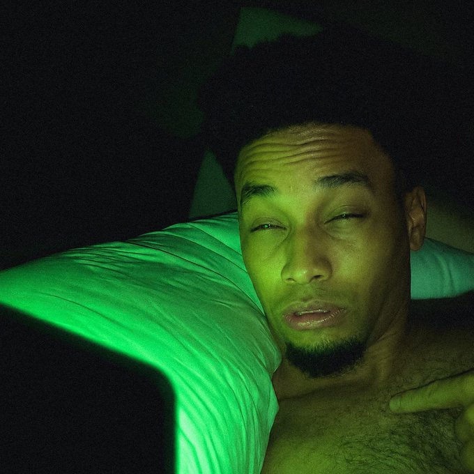 A close-up selfie of a young man with curly hair and a goatee in bed, illuminated by the bright green glow of a phone screen in a dark room.