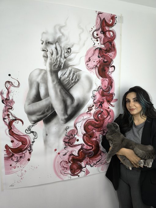 A woman with dark hair stands next to a large artistic poster featuring a black and white illustration of a man's torso with pink and red swirling decorative elements, while she holds a gray cat.