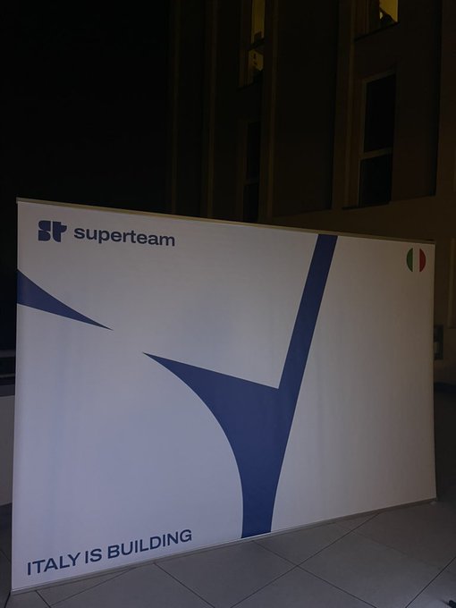 An illuminated 'superteam' sign at night displaying 'ITALY IS BUILDING' with a large stylized 'S' graphic and a small Italian flag.