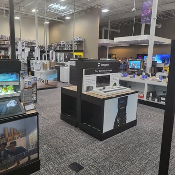 The image shows an electronics store with various electronic devices such as TVs and cell phones displayed throughout the space. The store also has a display of gaming consoles, including a Nintendo Wii console. A person can be seen browsing through the items on sale at this Best Buy. In addition to the electronics, there are two TVs in the scene - one is located near the center of the image while the other is positioned towards the right side. The store also features a large TV screen that displays information about the products available for purchase.