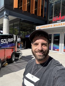 A man in a black cap and gray Solana t-shirt takes a selfie outside St. Lawrence Market with a Solana-branded truck behind him.
