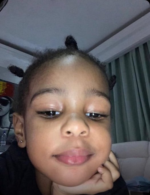 A close-up selfie of a young woman with her hair styled in small twists/space buns, wearing eyeliner, with curtains and a window in the background.