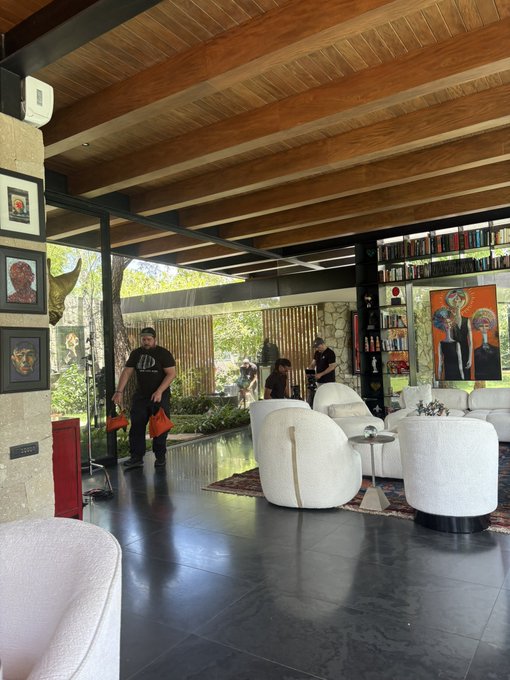 A man in a "D" shirt carries red shopping bags through a stylish mid-century modern home with vaulted wood ceilings, contemporary art, and white curved furniture. The scene appears to capture a casual visit, with the bag-laden guest contrasting the post's message about uncovering hidden stories and "chopping down" opponents.