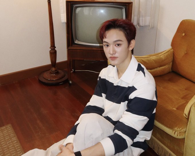 A young man with reddish-brown hair sits on the floor in a striped polo shirt, with a vintage TV and orange couch in the background.