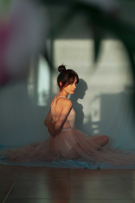 The image features a woman sitting cross-legged with her back against a wall, wearing a pink dress that appears to be made of tulle or chiffon fabric. The background includes a window and a plant, adding depth to the scene. The photo is titled "Good morning Whispered Glow" and has been shared on social media platforms.