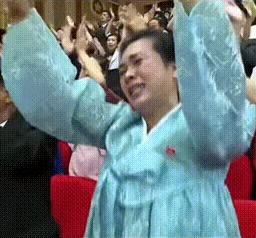 The image shows a man wearing traditional Korean clothing, possibly a blue dress or robe, standing up with his arms raised high above his head. The man appears to be in a joyful or celebratory mood, as he looks upwards towards something or someone. The background of the image is filled with people who are also dressed in similar attire and seem to be engaged in some sort of event or gathering.