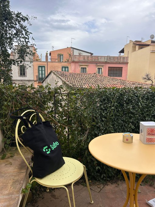 A small balcony scene features a yellow table and chair set with a black tote bag labeled 'Safe' and a candle, overlooking a hedge and Mediterranean-style buildings.