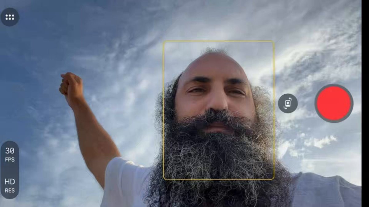 The image shows a man with a beard, wearing a white shirt, taking a selfie using his phone's front camera. The photo captures him from an overhead perspective, giving viewers a clear view of his facial features and the background behind him. The image also includes text elements that provide information about Bitcoin and other stock symbols.