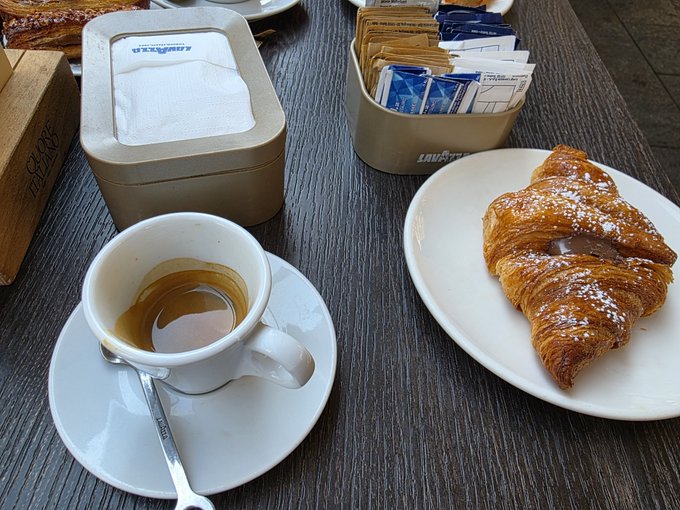An overhead view of a café table setting featuring a cup of black espresso on a saucer with a spoon, a chocolate-filled croissant dusted with powdered sugar on a plate, and two metal containers—one holding white napkins and the other filled with sugar packets.