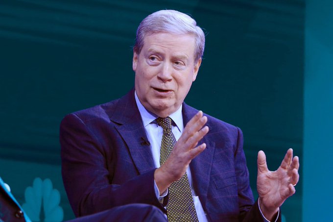 A man in a dark suit and patterned tie speaking while gesturing with his hands, photographed on a stage with NBC peacock logo visible in the background.