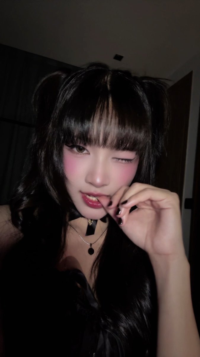 A close-up selfie of a young woman with dark hair in pigtails, heavy blush, and a black ribbon choker, winking at the camera while biting her lip.