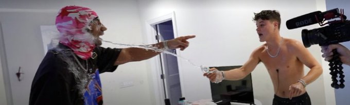 Two men face each other in what appears to be a video shoot, with one wearing a pink patterned head covering spraying liquid from his mouth while the other shirtless man holds a bottle, as a RØDE microphone is visible on the right.