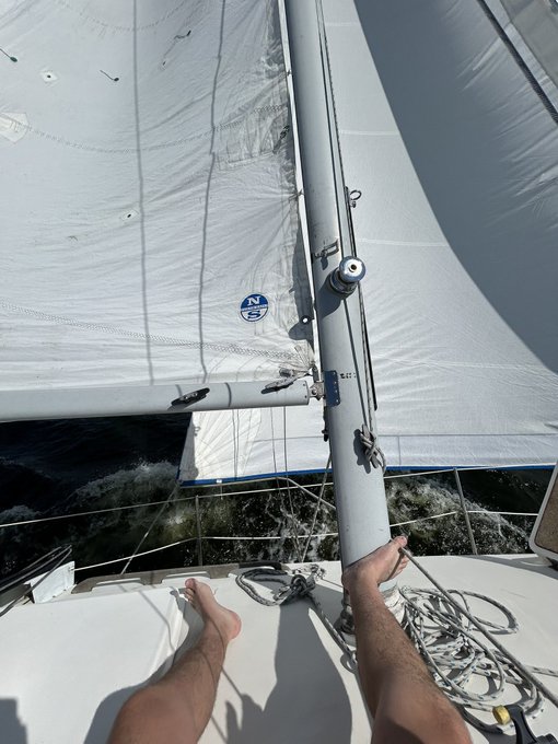 A first-person view from a sailboat shows a person's legs extended on the deck as they sail through choppy water.