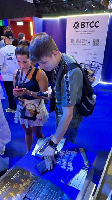 Two people stand at a booth labeled "BTCC" at a convention, with one young man reaching down to handle items on the display table.