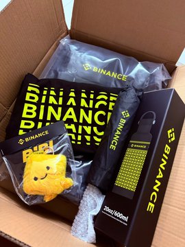 A cardboard box from Binance contains branded merchandise including a black umbrella, a "BINANCE" patterned pouch, a Pikachu figurine in plastic packaging, and what appears to be a 20ml/60ml product, all nestled in branded packaging materials.