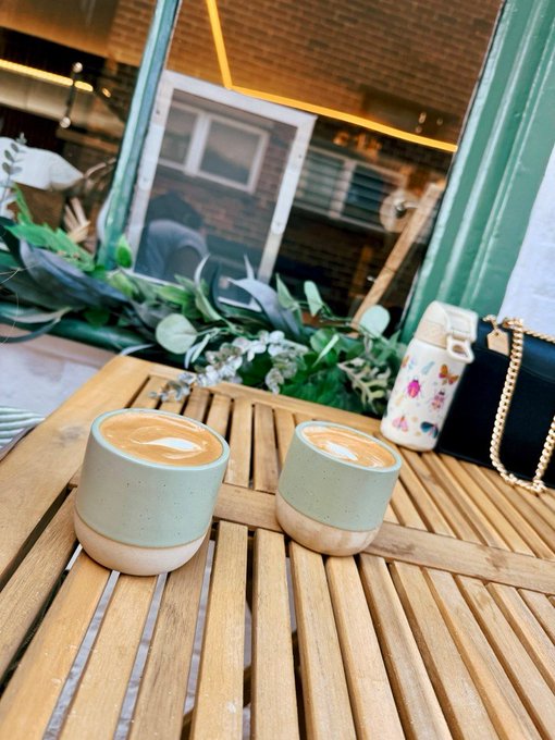 Two small ceramic cups of coffee with latte art sit on a slatted wooden table outside a cafe, with a black quilted handbag nearby.