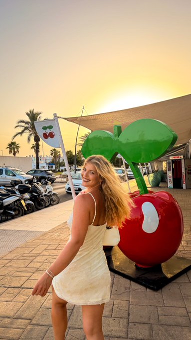 A blonde woman in a light yellow dress posing and smiling next to a large glossy red cherry sculpture with green leaves at an outdoor location during sunset.