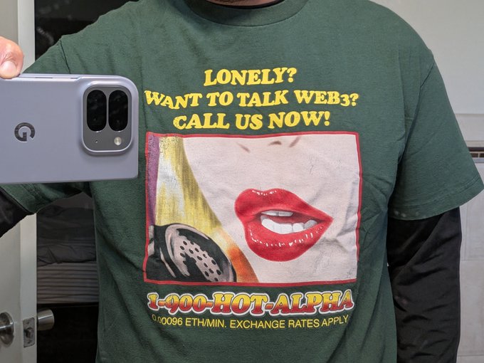 A person takes a mirror selfie wearing a green t-shirt that reads 'LONELY? WANT TO TALK WEB3? CALL US NOW! 1-400-HOT-ALPHA'.