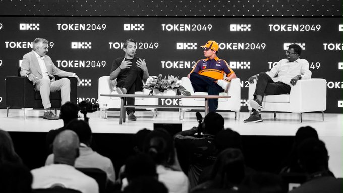 Four speakers, including F1 driver Lando Norris in his orange McLaren cap, participate in a panel discussion at the TOKEN2049 event, with the OKX and TOKEN2049 logos prominently displayed on the backdrop behind them. The image captures a moment on the OKX Main Stage where Norris joins industry experts to discuss how small, relentless improvements compound into a winning edge in both racing and financial markets.