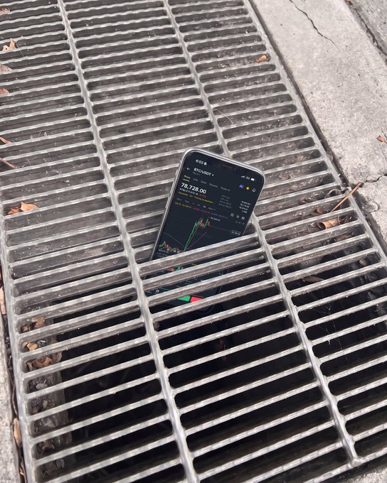 A smartphone displaying a Bitcoin (BTC) chart valued at $78,728 sits propped on a street grate, humorously illustrating the post's "if the chart loads, we're fine" sentiment—since the price is still refreshingly visible above ground rather than having "fallen through the cracks."