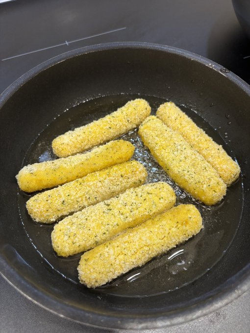 Breaded mozzarella sticks are arranged in a black frying pan with oil, ready to be cooked.