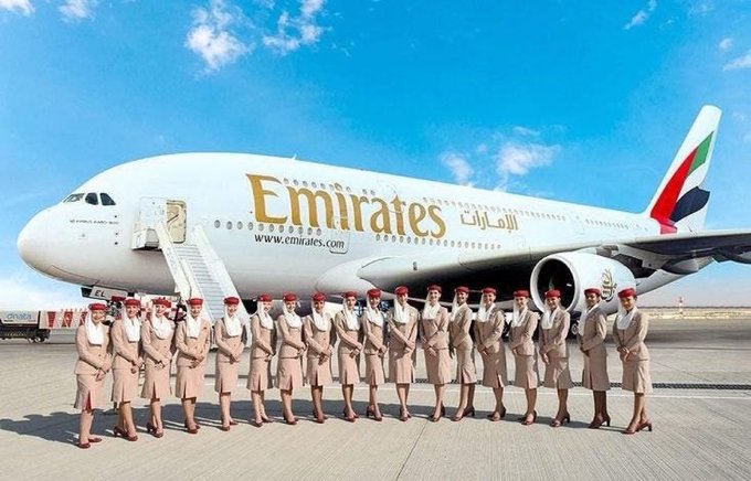A lineup of Emirates flight attendants in beige uniforms and red hats poses in front of a massive Emirates Airbus A380 jet, which displays the "Emirates" logo and "www.emirates.com" along its fuselage. The image visually reinforces the post's claim that the major airline is embracing cryptocurrency for flight bookings.