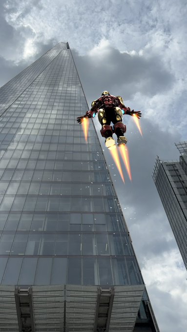 A low-angle shot of Iron Man in his Hulkbuster armor hovering with glowing rocket thrusters beside a tall modern glass skyscraper against a cloudy sky.