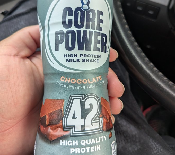 A hand holding a bottle of Core Power Chocolate High Protein Milk Shake, labeled '42g HIGH QUALITY PROTEIN,' photographed inside a car.