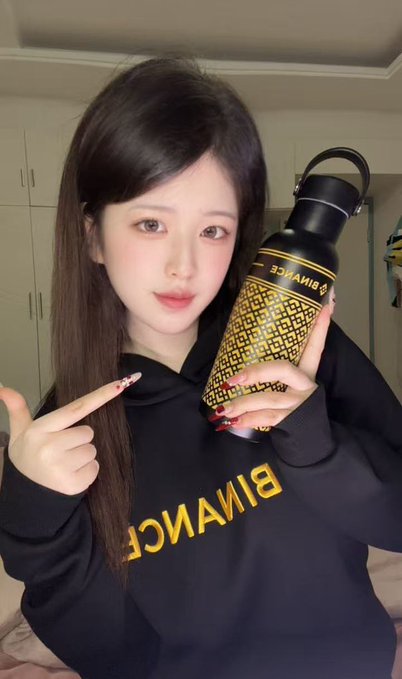 A young woman with long dark hair wearing a black hoodie points to a black and gold patterned Binance water bottle she is holding.
