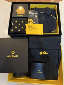 A Binance 9th Anniversary Gift Box containing a hoodie, "SWAG" socks, a yellow cap, and branded accessories, alongside a Binance Tennis Gift Set featuring a racket and matching apparel, all displayed in signature black and yellow Binance packaging.