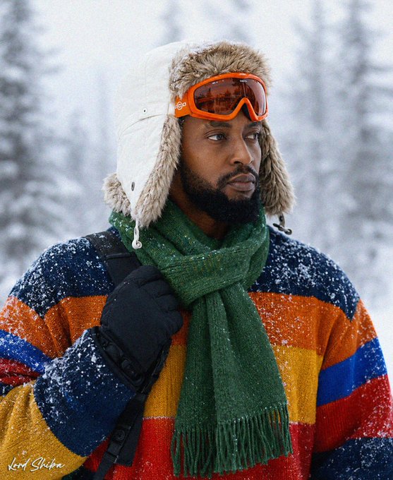 A bearded man stands in a snowy forest wearing a colorful striped sweater, a green scarf, black gloves, a fur-lined trapper hat, and orange ski goggles with the 'lord shirin' watermark in the corner.
