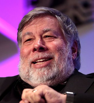 A photograph of Steve Wozniak, the Apple co-founder, smiling with a gray beard and wearing a black shirt against a purple background.