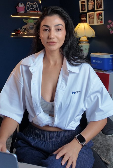 A woman poses in a home office setting wearing a partially unbuttoned white MEXC-branded shirt over a bralette, paired with a smartwatch and dark shorts, embodying a casual "corporate" look. The MEXC logo is visible on both her shirt and a box in the background, tying into the crypto exchange theme behind the caption "Feeling corporate today! Gm."