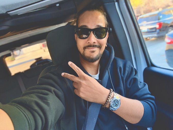 A man wearing black sunglasses and a dark hoodie takes a selfie in his car, flashing a peace sign while sporting a blue-faced watch. The relaxed, confident pose pairs perfectly with the casual "Gm CT" (Good Morning, Connecticut) greeting.