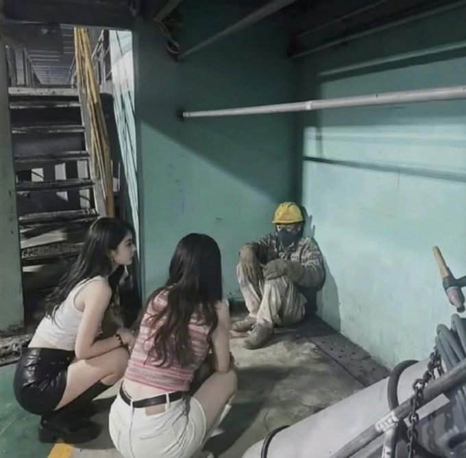 Two women crouch on the floor facing a worker wearing a yellow hard hat and mask who is seated against a wall in an industrial setting.