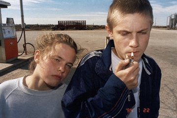A documentary-style photograph of two young people at what appears to be a gas station, with one smoking a cigarette.