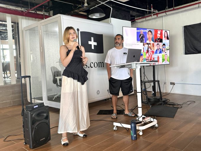 The image features a woman standing next to a man, both holding microphones as they appear to be giving a presentation or speech. They are surrounded by various electronic equipment, including laptops and speakers, suggesting that this might be an event related to technology or web3 development. The background includes a TV screen displaying multiple people's faces, indicating that the audience is engaged in the discussion.