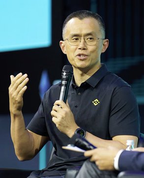 Changpeng Zhao (CZ), former CEO of Binance, speaking into a microphone while gesturing with his hand, wearing a dark polo shirt with the Binance logo and a smartwatch.