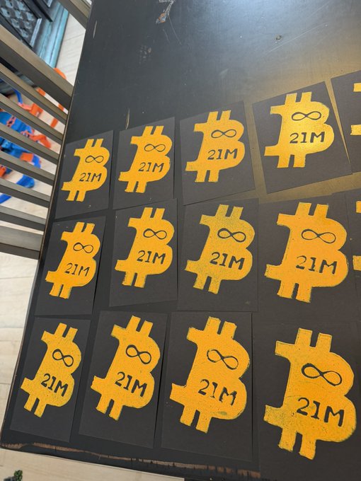 A photograph of multiple yellow Bitcoin '21M' stickers with an infinity symbol printed on them, arranged in rows on a dark surface, referencing Bitcoin's 21 million coin supply cap.